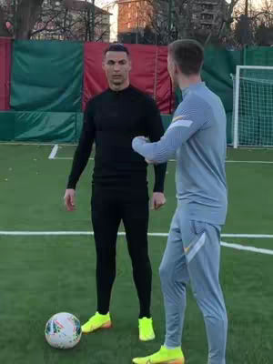 Two men standing on a green soccer field. One is formally dressed in a black long sleeve shirt and matching pants, while the other is casually dressed in a light blue athletic suit. A soccer ball is at the feet of the black clothed person.