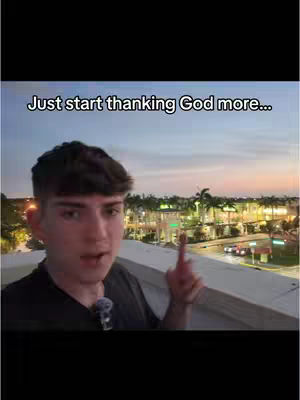 A young man with short dark hair is centered in the frame, pointing upwards with his right index finger. He's wearing a dark t-shirt and a small microphone is clipped to it. He's standing on a rooftop, with a city view in the background at dusk, various illuminated buildings and palm trees are visible, and the sky displays colors of the sunset. The text overlay at the top declares "Just start thanking God more..."