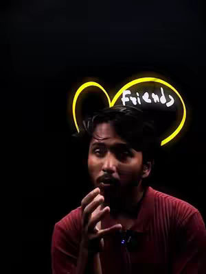 A man with dark hair and a red shirt is speaking, with a blurred yellow heart shape behind his head with the word 'friends' in it, on a black backdrop. His hand gestures convey emotion or emphasis. The lighting seems dramatic.