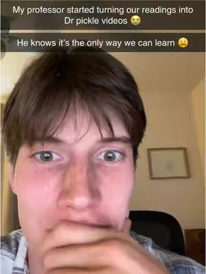 A selfie video shows a young man with brown hair looking directly at the camera. He has a slightly surprised expression, with his hand near his face. The background is a cluttered room with a framed picture on the wall. Text overlays at the top state, "My professor started turning our readings into Dr pickle videos", followed by, "He knows it's the only way we can learn".