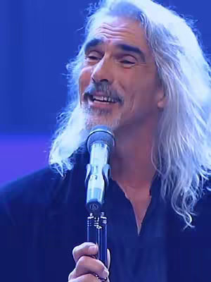 A man with long white hair and a mustache is singing into a microphone. He is wearing a black shirt and the background is blue.