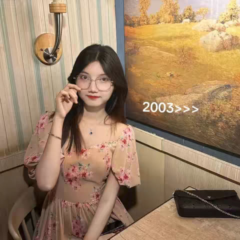 A young woman with long dark hair and glasses sits at a table in a restaurant-like setting. She wears a floral dress and looks toward the camera, holding her glasses with a slight smile. The background features a painting and a wall sconce.