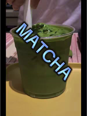 A close-up shot of a clear plastic cup filled with matcha. The matcha, a vibrant green, is topped with a layer of foam. The word 'MATCHA' is overlaid on the image in a blue, outlined font. A hand is visible in the background and a white straw is in the cup.