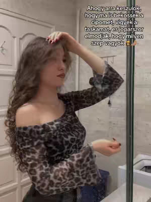 A woman with long curly hair stands in front of a mirror, angled slightly to the side. She wears an off-the-shoulder top with a leopard print pattern. She has her right arm raised, touching her hair. Her left arm is down, with her hand visible. The background shows a light-colored door or wall and a bathroom mirror and a toilet.