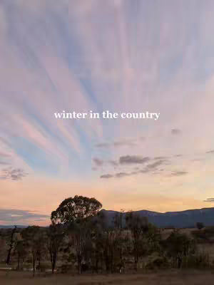 An image of a dusky sky and a rural scene. The sky is filled with streaks of pink and blue from the setting sun or rising dawn. Silhouetted against the horizon are trees and hills. In the center of the image, the words "winter in the country" are written in a simple, white font.