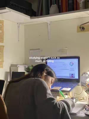 A person is sitting at a desk, facing away from the camera, with their head down and focused on writing. A laptop sits open on the desk, displaying a screen with an interface. The background includes a shelf with books and a small decorative item, along with items like a desk lamp and some plants. The color scheme is warm, with beige-painted walls and warm lighting.