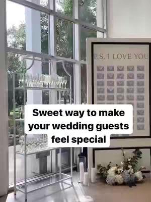 The cover image features a well-lit setting, likely indoors, with a large window dominating the background. The window allows natural light to flood the scene, revealing a glimpse of trees and greenery outside. In the foreground, there's a silver metallic shelving unit displaying glassware, suggesting a bar or drink station. To the right, a framed artwork or sign, reading "P.S. I LOVE YOU", adds a decorative element. Further down, a vase of flowers sits on a table or surface, along with some candles. Overlayed on the scene is white text in a rectangle, stating "Sweet way to make your wedding guests feel special".