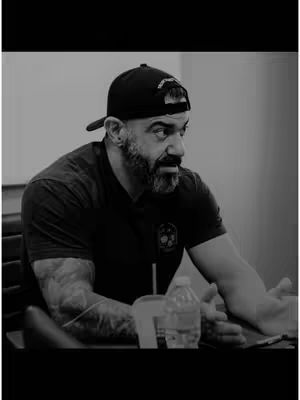 Black and white photo of a man wearing a black cap and shirt, sitting at a table with a water bottle and a drink, with a thoughtful expression.