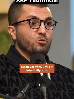 A man with short, dark hair and glasses is speaking. There is a microphone in front of him and a text overlay at the top and bottom of the frame.