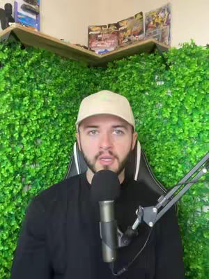 A medium shot shows a man in front of an artificial green wall. He is wearing a black shirt and a beige baseball cap. He is sitting in a gaming chair and talking. In front of him is a microphone.