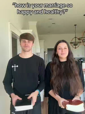 A couple, a man and a woman, stand side-by-side holding books while looking directly at the camera in a bright, indoor setting. The man wears a black shirt with a cross symbol, and the woman also wears black. The background features a neutral wall, a door frame, and hints of a kitchen setting.