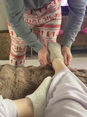 A person is lying in bed, viewed from a low angle. Another person, whose body from the knees up is visible, is standing over them, with their hands clasped on the lying person's feet, The person standing is wearing patterned pajama pants with red and white decorative shapes, and a gray long-sleeved top. The person in bed appears to be wearing light-colored socks.