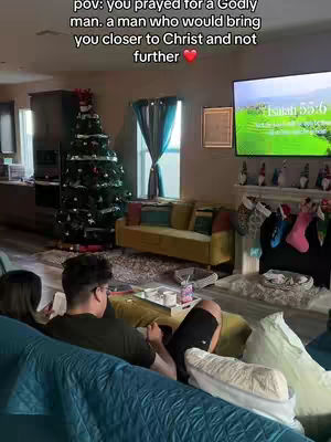 A couple is relaxing in a cozy living room decorated for Christmas. The man is sitting with his back to the camera, and the woman is to the left, with a Christmas tree in the background.