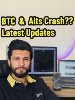 A man with dark hair and a beard is centered in front of a desk with a computer monitor and a laptop showing a chart. The overlay text is yellow with a black stroke stating, “BTC & Alts Crash?? Latest Updates”. The background has a soft purple gradient.