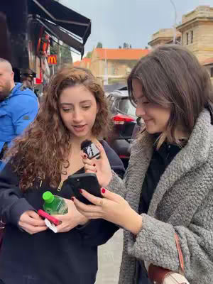 Two young women are standing on a city street, one holding a phone and microphone. The woman on the left is holding her phone and other items. Behind them, there are building and a car. The overall tone of the image is casual and modern.