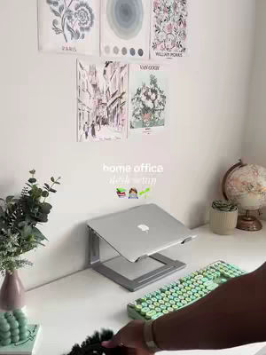 A home office or guest room setting with a white desk, a laptop on a stand, and a colorful mechanical keyboard, with decorative posters on the white wall and a globe on the right.