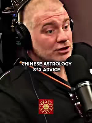 A man with short, light hair and a beard is centered in the shot, wearing a black jacket and headphones. A microphone is positioned near his mouth. The background is dark, with a red decorative element at the bottom center which shows a symbol. Text overlayed near the top of the image reads 'CHINESE ASTROLOGY S*X ADVICE'. The composition is a medium close-up, focusing on the man's face and upper body.