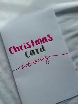 A white piece of paper rests on a soft, white surface, possibly a bed. Bold text in pink and green colors announces the video topic: 'Christmas card ideas'.