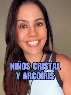 A smiling woman with dark hair is centered in the frame. She wears a light-colored tank top with a navy blue trim. A text overlay in blue and white, 'NIÑOS CRISTAL Y ARCOÍRIS' (Crystal and Rainbow Children) is at the bottom of the frame. The background is plain and neutral.