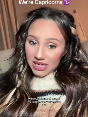 A woman with long wavy brown hair is centered in a close-up shot of her face. She has pink blush and lipstick, and is looking directly at the camera with her mouth slightly open. Above her head, the words 'We're Capricorns' are visible, accompanied by a Capricorn symbol. The background appears to be a dimly lit room, and she's wearing a white sweater. The overall composition is focused on her expressive face.