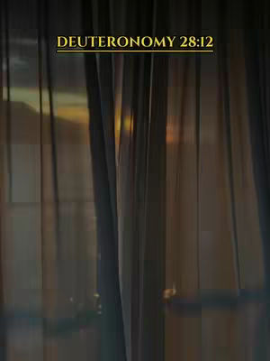 The image showcases a curtain partially obscuring a view of a sunset. A gold-colored rectangular text overlay at the top states "DEUTERONOMY 28:12". The curtains, likely sheer, are vertical and arranged in a way that allows a glimpse of a warm, golden light or the setting sun in the background. The composition is simple, with a warm color palette.