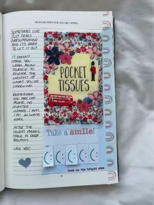 A journal spread with handwritten text and various decorative elements. In the center, a small square is adorned with floral patterns and the words "POCKET TISSUES." Below this, another square contains cut-out smileys, and text that reads, "Take a smile!" and "look on the bright side." The journal page is open, featuring lined paper with more handwritten text on the left, a blue heart drawn in the corner and rainbow-colored decorative tape along the right edge.