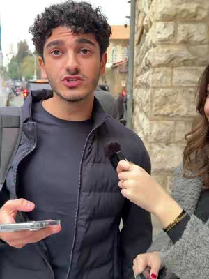 A man with curly dark hair is centered, facing the camera with a concerned expression. He is wearing a black puffer jacket over a black shirt. A person's hand with a microphone is on the right of the man and another person is partly visible from the right side. The background shows a building with stone blocks and a glimpse of a busy street.