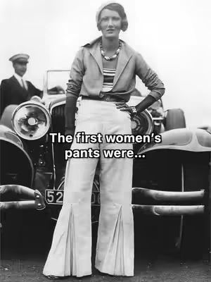 A black and white photo showcasing a woman standing in front of an antique car. She's wearing 1930s-style attire: a tailored jacket, wide-legged pants, and a cloche hat. A man in a suit and cap stands in the background. The text overlay reads: "The first women's pants were..."