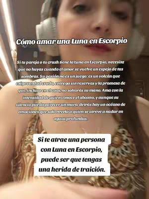 A selfie-style video cover image shows a woman with headphones around her neck, making direct eye contact with the viewer. Text overlays are placed on the image, with the main header in bold white font reading 'Cómo amar una Luna en Escorpio', positioned at the top. The woman is fair-skinned and appears to be indoors, with some lighting visible in the background.