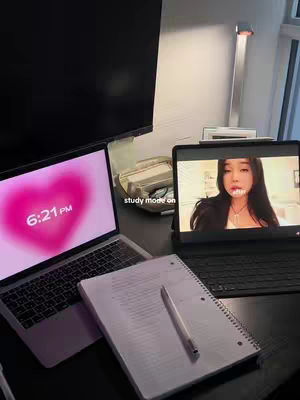 A well-lit image shows a study setup. Two open laptops are positioned on a desk. The laptop on the left displays a pink heart-shaped graphic with the time. A notebook and a pen are placed in the foreground. A tablet shows of a person and text. A monitor and a desk lamp is visible on the background