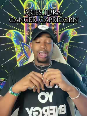 A man with dark skin is centered in the frame, wearing a black t-shirt printed with white text, a black baseball cap, and silver jewelry. He has a slight smile and is looking directly at the camera. Behind him is a colorful tapestry with an intricate design featuring hands and stars.