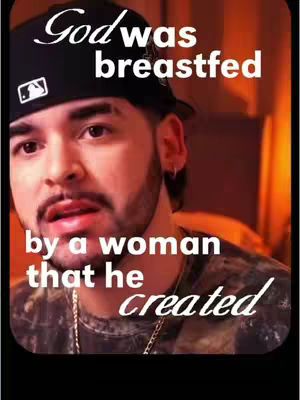 A man wearing a black cap with white text and a camo shirt is the main subject of the image. Above his head, the overlay reads 'God was breastfed' in a stylish, somewhat cursive font. Below, in the same font, it says 'by a woman that he created.' The man has a beard and is looking intently at the camera, with a slight expression of surprise or contemplation. The lighting is warm, with a focus on the face against a blurred background, enhancing the contrast and drawing attention to the message.