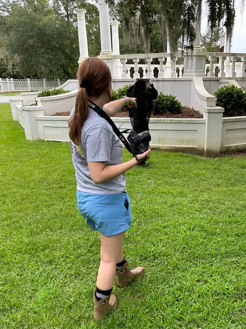 A woman with red hair in a low ponytail, wearing a gray t-shirt and blue shorts, holds a camera. She's pointing towards a person obscured by a black object, with a white structure in the background.