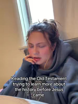 A young woman with light skin and a messy bun hairstyle is shown reading. She's wearing a dark blue hoodie with the hood down, and her expression suggests deep concentration as she slightly bites her lip. The background is a warm-toned room with a window visible on the left.
