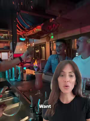 A woman is in the foreground, with a bar and people in the background at a restaurant. She is looking directly at the camera and has a word bubble with "Want" overlaid. Background elements include a lit bar, other people, and a bartender preparing a drink at the bar.