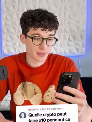 A young man with curly dark hair and glasses is in a selfie-style video. He wears a red t-shirt featuring a teddy bear design and holds a black smartphone. Behind him, a textured white wall provides a simple backdrop. A comment bubble at the bottom displays French text about crypto.