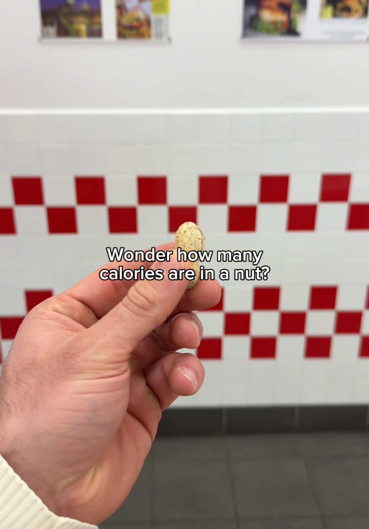 A hand is holding a peanut in front of a tiled wall. The wall is white with a grid of red squares. The hand is pale and appears to be in a kitchen or dining area. The text overlay asks about the number of calories in a nut.