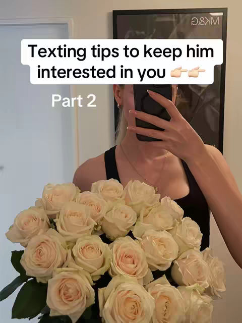 A woman in a black top is holding a phone in front of her face, likely taking a selfie in a mirror. She's holding a large bouquet of white roses. The text overlay reads, 'Texting tips to keep him interested in you.'