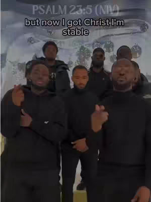 The image shows a group of Black men, likely in their 20s or 30s, posed together. The background is a light-colored, possibly digitally enhanced, design, which is mostly out of focus except for a blurred pattern. The men are dressed in black hoodies and are gesturing with their hands as if they are singing or communicating. The overlay text is set in a white, bold typeface on the top of the screen.