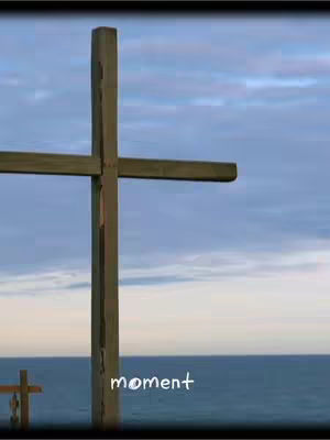A wooden cross is set against a backdrop of a cloudy blue sky and the ocean in the distance. The cross occupies the center of the frame, with the ocean horizon visible at the bottom and a slightly darker black border around the image.