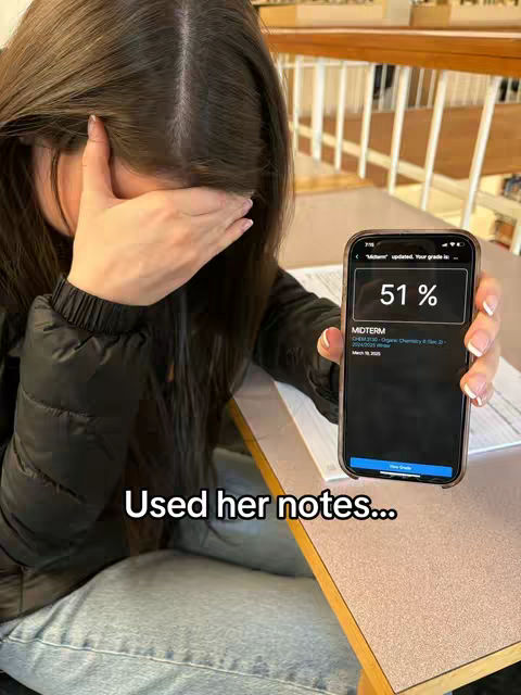 A young woman is sitting at a desk in a library, holding a phone displaying a low grade (51%) over her face. Her other hand is raised to her face in a gesture of frustration or disappointment. The background includes bookshelves and desks, indicating a study environment.