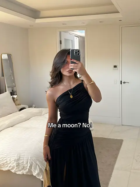 A woman in a black one-shoulder dress is taking a mirror selfie. The room has a neutral palette with light-colored walls, a beige rug, and white bedding. Text overlayed on the image reads, 'Me a moon? No!'