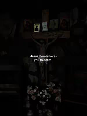 The image is a dimly lit, vertical shot focused on a crucifix statue. A figure of Jesus Christ is depicted on the cross, positioned centrally. Below the cross, there is a collection of flowers, possibly roses, in a close arrangement. Above the crossbar, various framed pictures or icons are displayed. The background is dark, suggesting a church setting.