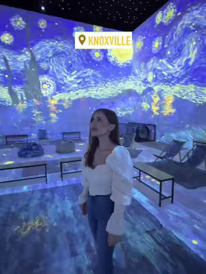 The cover image shows a woman standing in a room that has been transformed using projection mapping. The walls and ceiling are covered in a swirling blue and yellow pattern reminiscent of Van Gogh's "Starry Night." The woman is facing the camera, wearing a white top with puffy sleeves and blue jeans. A white rectangle with "KNOXVILLE" and a location pin is overlayed the image. The overall impression is one of art and immersive experience.