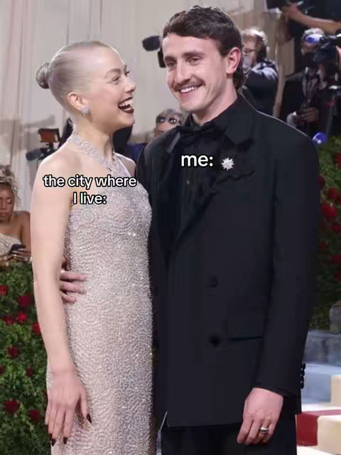 The cover image features a man and a woman embracing and laughing at an event, posing in front of a red carpet and a lush green backdrop. The woman is on the left, wearing a shimmery, silver dress, with her hair styled in a bun. The man is on the right, wearing a tuxedo with a bow tie and a dark jacket, and has a mustache. Text overlays read 'the city where I live:' on the woman's side and 'me:' on the man's side.
