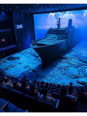 A view from inside a movie theater, looking at a large screen depicting a shipwreck on the ocean floor. The audience is seated in dark blue chairs, looking up at the screen. The screen shows a detailed scene of a sunken ship resting on the seabed with a blue hued filter, surrounded by water. The theater has a modern design, with black walls and a minimalist aesthetic.
