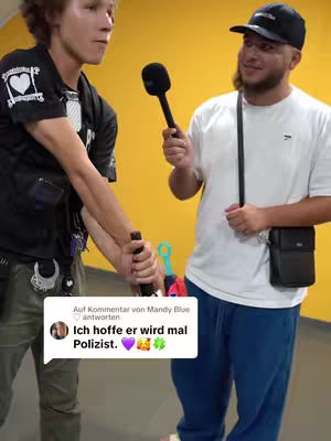 Two men are featured in an interview setting. The man on the left is wearing a black shirt with patches on the shoulders, and the man on the right is wearing a white t-shirt and a black cap. They are standing in front of a yellow wall.