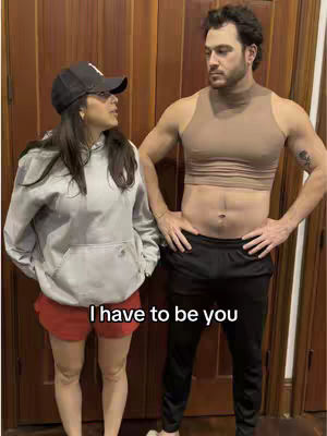 A woman and a man are standing side by side, facing each other, in front of a wood-paneled wall. The woman on the left is wearing a black cap, a gray hoodie, and short red shorts. The man on the right is wearing a tan crop top and black sweatpants. The text "I have to be you" is overlaid at the bottom of the image.