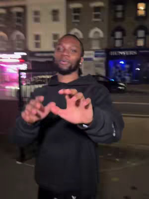 A man wearing a black hoodie stands on a city street at night. He gestures with his hands, seemingly talking while looking off-camera. Buildings and a street are visible in the background, with some neon signs illuminating the scene.
