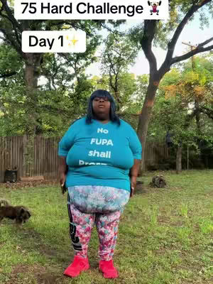 A full-body shot of a person standing outdoors. The person, who appears to be a woman, is wearing a blue t-shirt with the words "No FUPA shall DRESS" printed across it. She is also wearing colorful patterned leggings and bright pink shoes. The background consists of a grassy area with trees and foliage, giving it a natural setting.